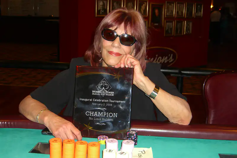 Woman in sunglasses at a poker table holding a champion trophy behind stacks of chips